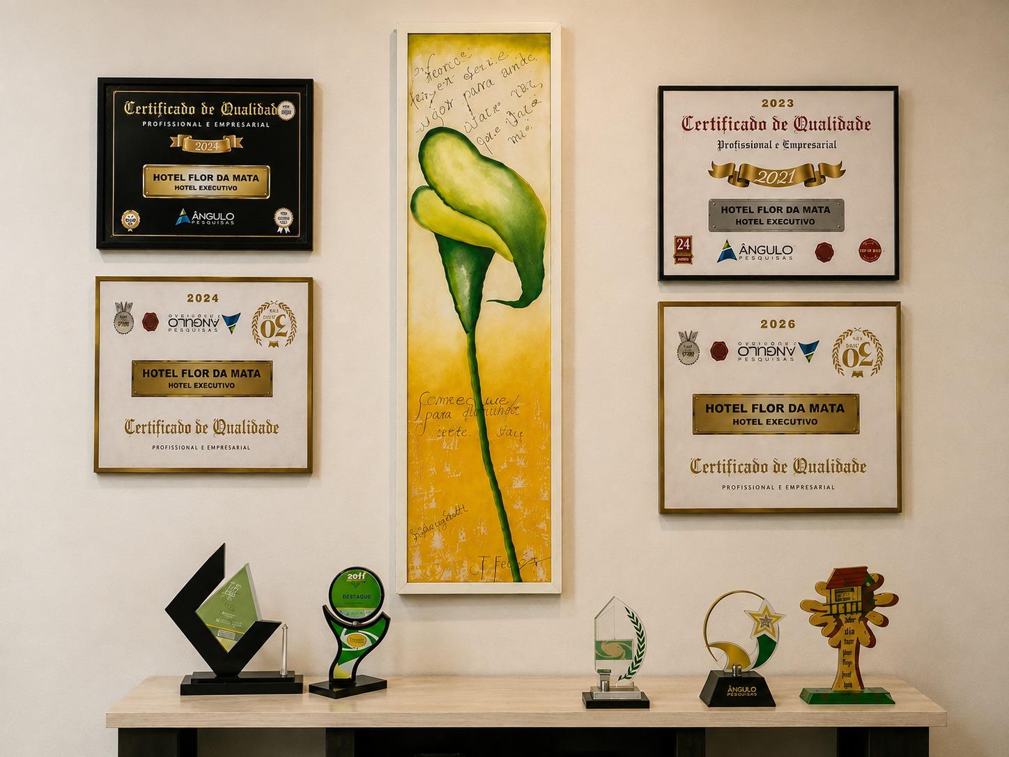 Parede do Hotel Flor da Mata com quatro Certificados de Qualidade Profissional e Empresarial da Ângulo Pesquisas, quadro de uma flor amarela ao centro e troféus sobre o aparador